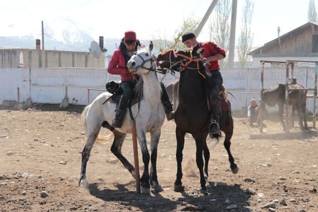 Deux hommes à cheval se font face dans un paddock poussiéreux avec des montagnes enneigées visibles au loin.