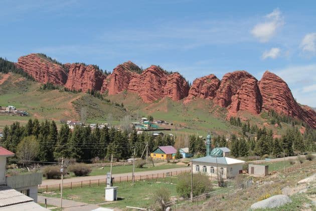 Un petit village avec une mosquée se trouve au pied d'une grande chaîne de montagnes rocheuses rouges et stratifiées sous un ciel bleu.