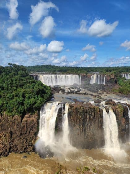 Una vista panoramica di imponenti cascate che scendono da scogliere rocciose, con una giungla rigogliosa sulle sponde sotto un cielo azzurro nuvoloso.