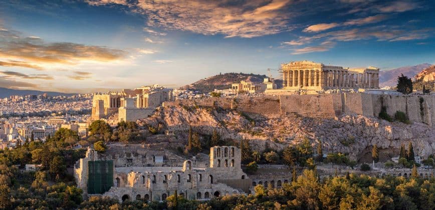 A historic acropolis with ancient ruins sits on a rocky hill overlooking a sprawling city under a colorful sunset sky.