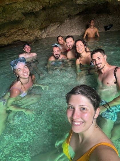 A WeRoad group trip smiling for a selfie while swimming in the clear, green water of a rocky sea cave.