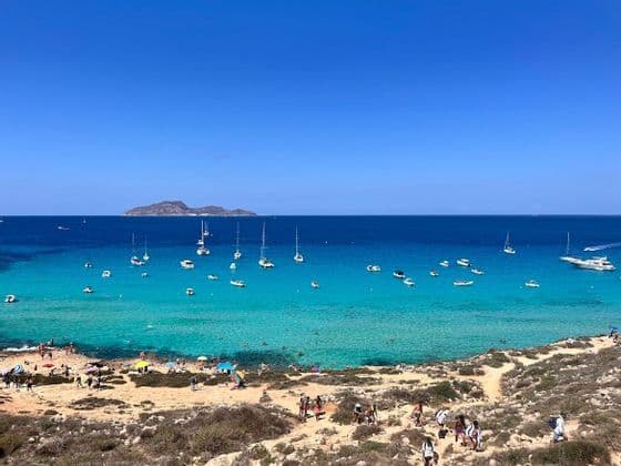 A WeRoad group trip walks on a coastal path overlooking a sunny bay with turquoise water, many boats, and a distant island.