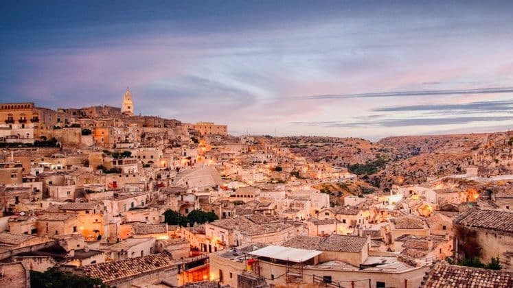 Un borgo di antichi edifici in pietra e un campanile incastonato nella collina, illuminato al crepuscolo accanto a un burrone.