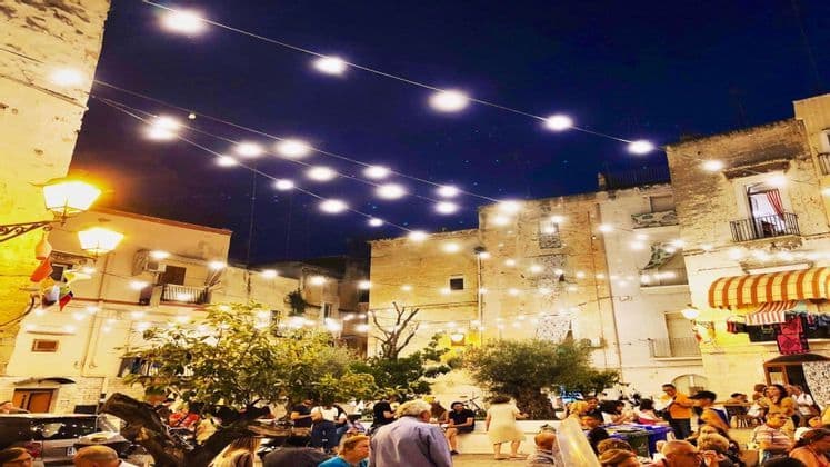 Una folla di persone si è radunata in una piazza di notte, illuminata da file di luci appese tra vecchi edifici.