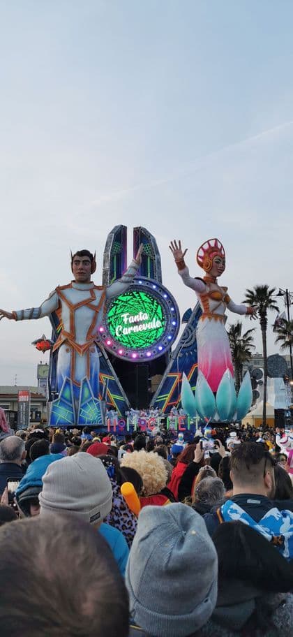 Una grande folla osserva un carro allegorico di carnevale con due figure futuristiche giganti sotto un cielo coperto.