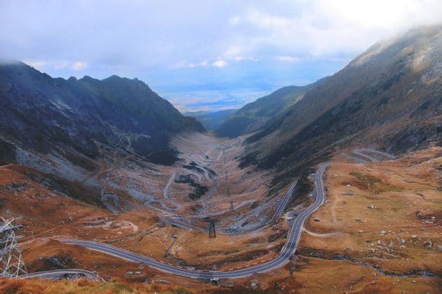 Vue aérienne d'une route sinueuse avec de nombreux virages en épingle, descendant dans une vaste vallée montagneuse sous un ciel nuageux.