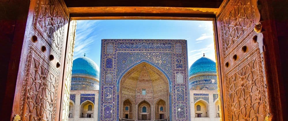 Una moschea ornata con cupole blu e un grande ingresso ad arco, vista attraverso due porte di legno aperte e finemente intagliate.