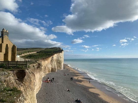 Una vista costiera di persone su una spiaggia di ciottoli ai piedi di scogliere bianche sotto un cielo azzurro con nuvole.