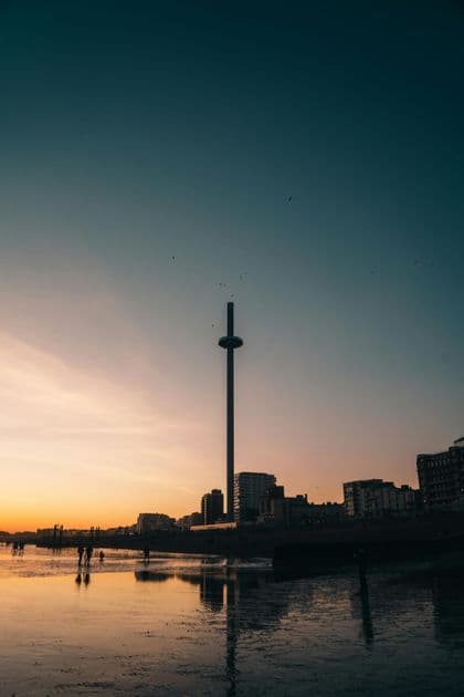 Un'alta torre panoramica e uno skyline cittadino si stagliano contro un tramonto, con i loro riflessi visibili sulla spiaggia bagnata sottostante.