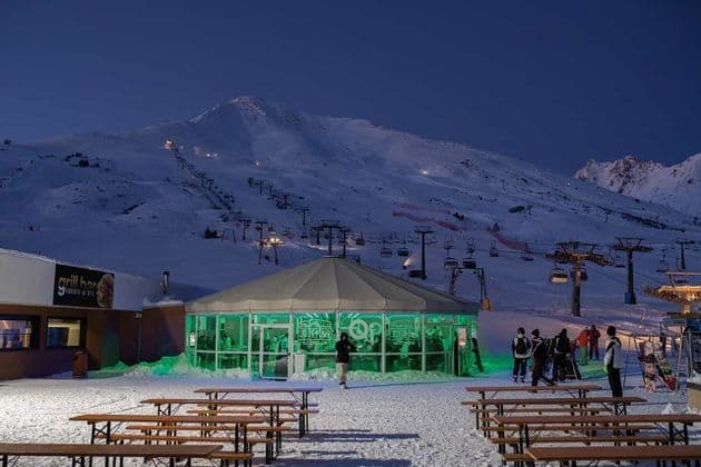 Un bar circolare illuminato di verde in una località sciistica di notte, con una montagna innevata e impianti di risalita sullo sfondo.