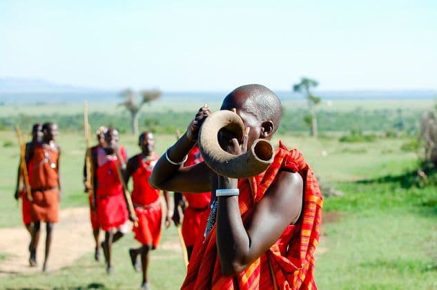 A Maasai man in red traditional clothing blows a horn while other tribesmen walk behind him in a grassy savanna.