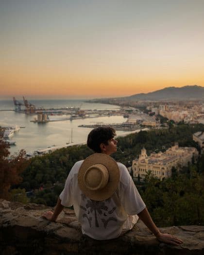 A person with a straw hat on their back sits on a stone wall, looking out over a coastal city and harbor at sunset.