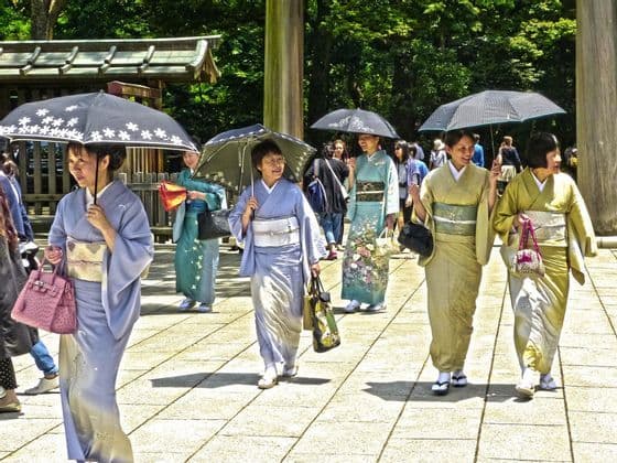 Un gruppo di donne giapponesi in kimono colorati cammina su un sentiero lastricato, tenendo ombrelli aperti per l'ombra.