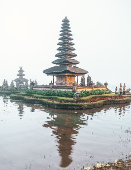 Un tempio tradizionale a più livelli sorge su una piccola isola, il cui riflesso si specchia sull'acqua calma sotto un cielo nebbioso.