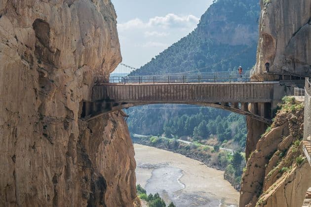 A high bridge connects two sheer rock cliffs over a wide river in a deep canyon, with two people standing on it.