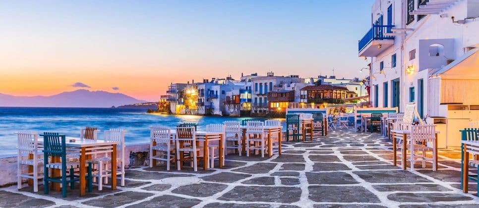 Patio in pietra di un ristorante sul mare con tavoli vuoti e vista su una località costiera con edifici bianchi al tramonto.