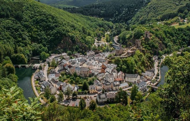 Una vista aerea di un piccolo villaggio annidato in un'ansa di fiume, con un castello in rovina su una collina, circondato da verdi montagne boscose.