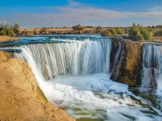 Un'ampia cascata scende da una scogliera rocciosa in un fiume impetuoso, con un paesaggio arido sullo sfondo sotto un cielo azzurro.