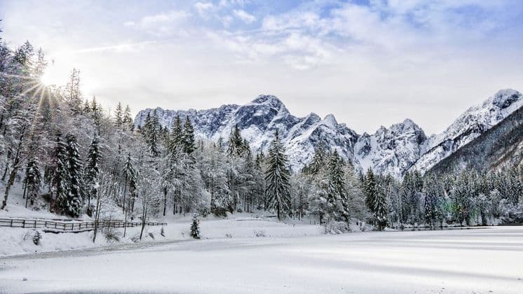 La luce del sole splende su una pineta coperta di neve e un lago ghiacciato, con una catena montuosa scoscesa sullo sfondo.