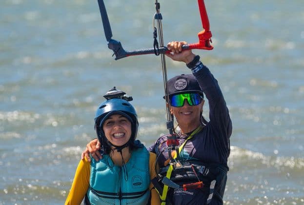 Due donne in attrezzatura per sport acquatici, inclusi caschi e giubbotti di salvataggio, sorridono nell'acqua mentre una tiene in mano una barra di controllo per kitesurf.