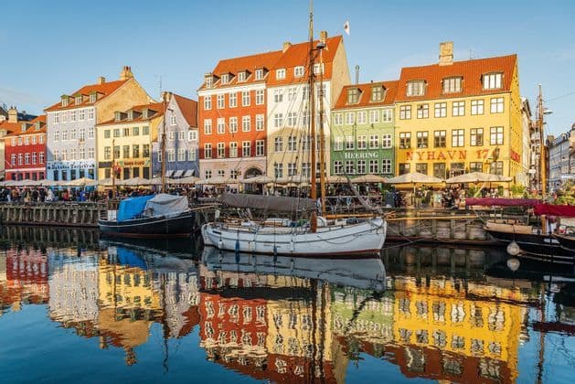 Colorful buildings and moored sailboats are reflected in the calm water of a canal under a clear blue sky.
