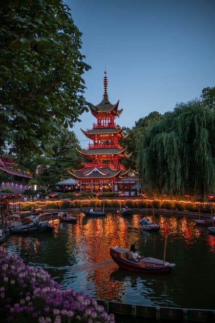A large, red pagoda is illuminated at twilight, its lights reflecting on a pond where people ride in small boats.