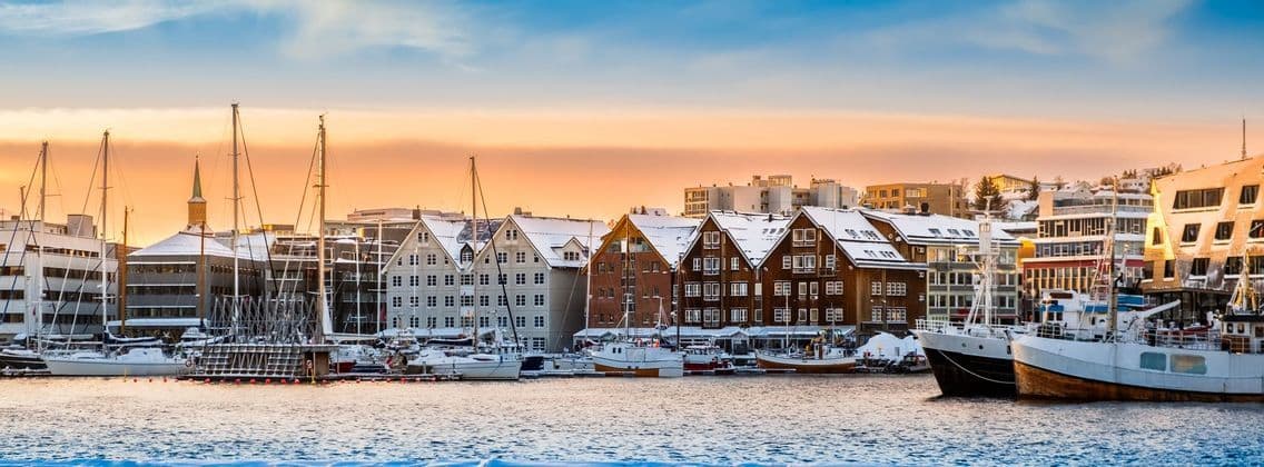 Un panorama di un porto innevato con barche ormeggiate lungo un lungomare costeggiato da edifici tradizionali al tramonto.