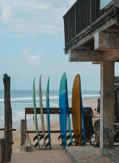 Tablas de surf coloridas en un estante de madera en una playa de arena, con olas del océano visibles al fondo.