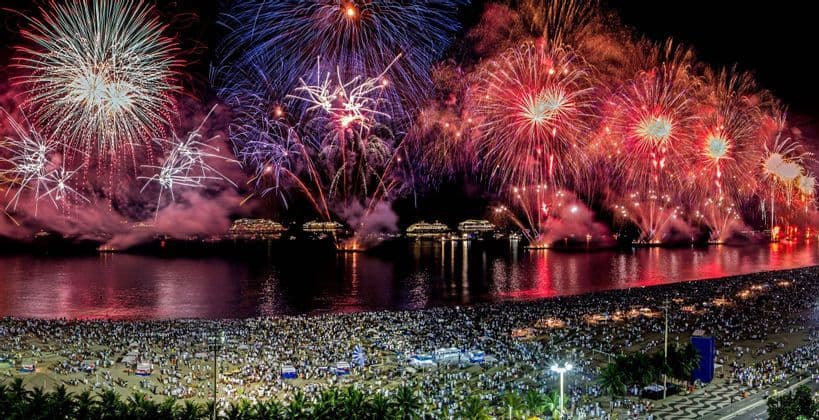 Un enorme spettacolo di fuochi d'artificio illumina il cielo notturno sopra una spiaggia affollata di persone che osservano dalla riva.