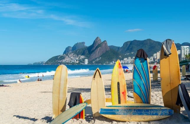 Una fila di tavole da surf colorate si trova sulla sabbia di una spiaggia, con l'oceano, una città e le montagne sullo sfondo.