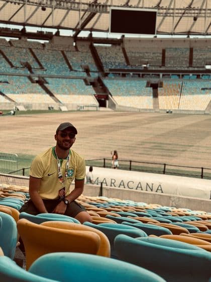 Un uomo sorridente in maglia gialla e occhiali da sole siede sui sedili colorati di uno stadio vuoto.