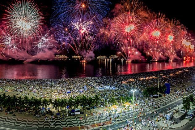 Coloridos fuegos artificiales estallan en el cielo nocturno sobre una bahía con barcos, contemplados por una multitud masiva en una playa de arena.