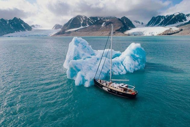 Un viaggio di gruppo WeRoad in barca a vela naviga accanto a un grande iceberg in acque artiche, con montagne e ghiacciai sullo sfondo.