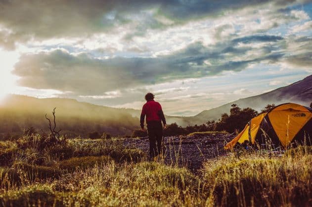 A person in a red jacket stands beside a yellow tent in a mountain valley, watching the sunrise through the clouds.