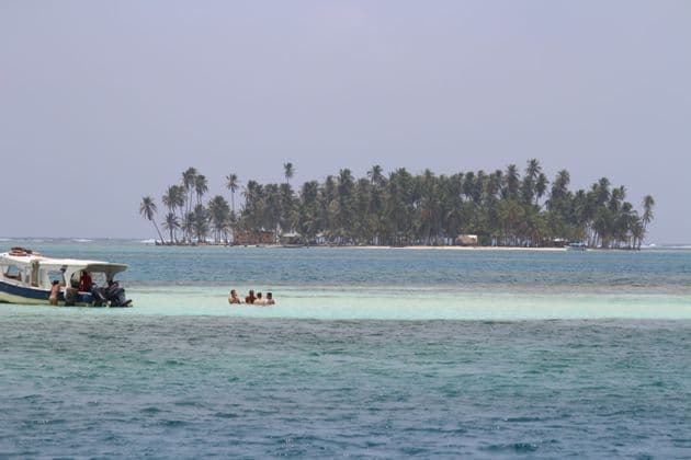 Un groupe WeRoad se détend dans l'eau turquoise peu profonde près d'un bateau, avec une île tropicale couverte de palmiers en arrière-plan.