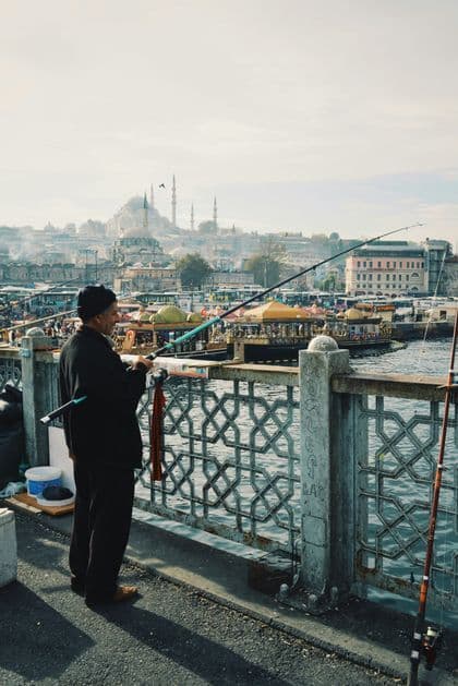Un hombre con gorro negro pesca desde un puente, con el horizonte de la ciudad y una gran mezquita visibles a la distancia.