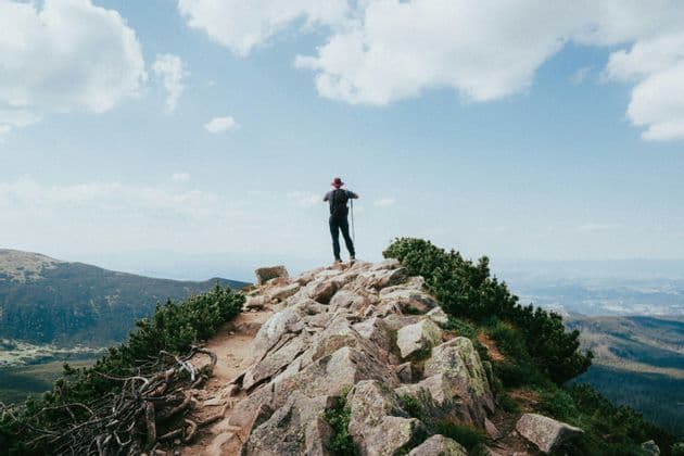 Ein Wanderer mit Rucksack und Trekkingstock steht auf einem felsigen Berggipfel und blickt auf eine weite Landschaft unter blauem Himmel mit Wolken.