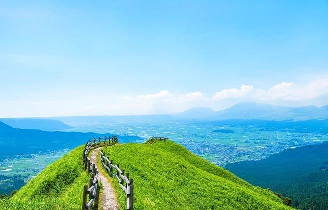 Un sentiero sterrato tortuoso con una staccionata di legno su una collina di un verde brillante che si affaccia su una valle e montagne distanti sotto un cielo blu.