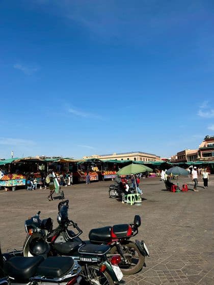 Motos aparcadas en primer plano de un bullicioso mercado al aire libre con puestos de fruta en una gran plaza bajo un cielo azul claro.