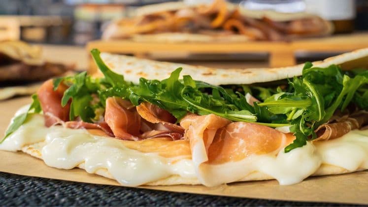 Primo piano di una piadina piegata ripiena di prosciutto, formaggio fuso e rucola fresca su carta marrone.