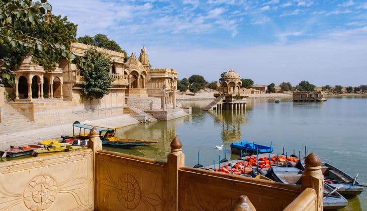 Ornate sandstone temples and pavilions sit on the edge of a lake, with colorful boats docked along the stone steps under a blue sky.