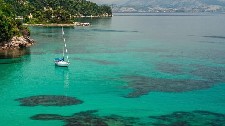Una barca a vela è ancorata in una baia di acqua cristallina turchese vicino a una costa verde lussureggiante.