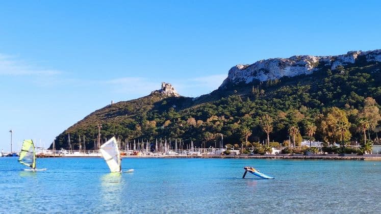 Windsurfistas en aguas tranquilas y azules en una bahía con un puerto deportivo al pie de una montaña verde y rocosa bajo un cielo despejado.