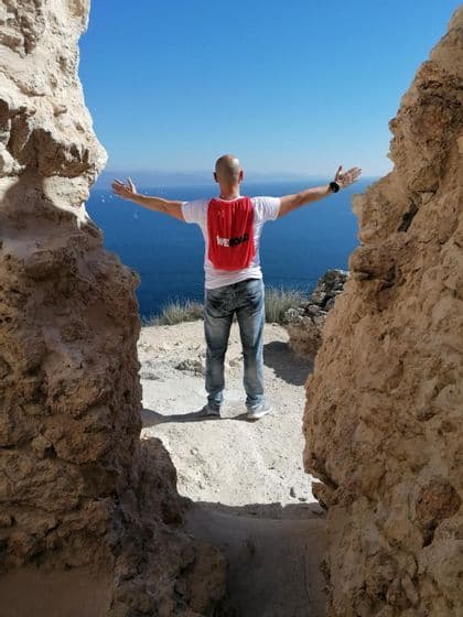 Un hombre con una mochila WeRoad roja está de pie con los brazos abiertos en un acantilado, contemplando el mar azul y los veleros.