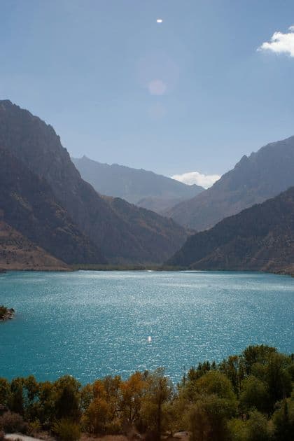 Un lago turchese brilla al sole, incorniciato da ripide montagne rocciose e alberi in primo piano, sotto un cielo azzurro.