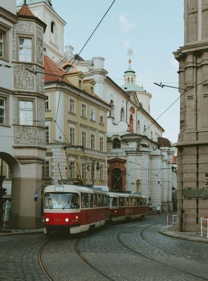 Un tram rosso e bianco percorre i binari lungo una via acciottolata e tortuosa, fiancheggiata da edifici storici e decorati.