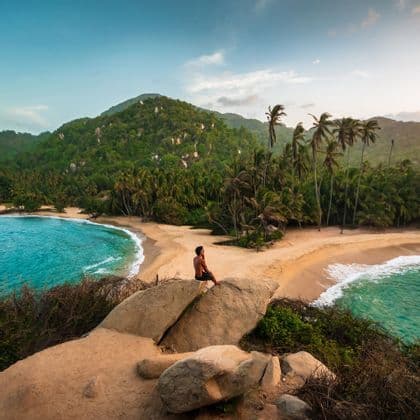 Ein Mann sitzt auf einem großen Felsen und überblickt zwei tropische Strände mit türkisfarbenem Wasser, Palmen und einem üppig grünen Hügel.