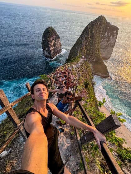 Un homme prend un selfie lors d'un voyage de groupe WeRoad sur un chemin étroit à flanc de falaise surplombant une plage et l'océan turquoise au coucher du soleil.
