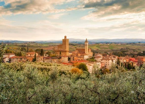 Un borgo con una torre in pietra e una chiesa è immerso tra dolci colline verdi, con ulivi in primo piano.