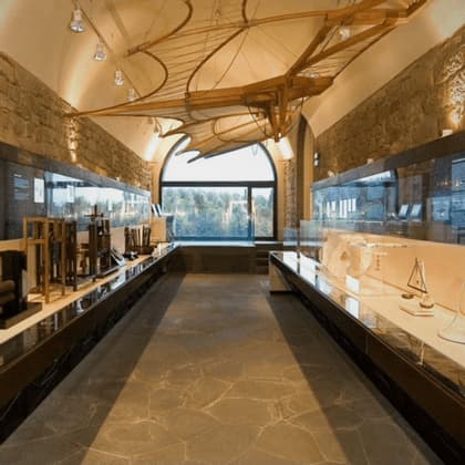 Un grande modello in legno di una macchina volante storica è appeso al soffitto di una galleria museale con pareti in pietra e teche di vetro.